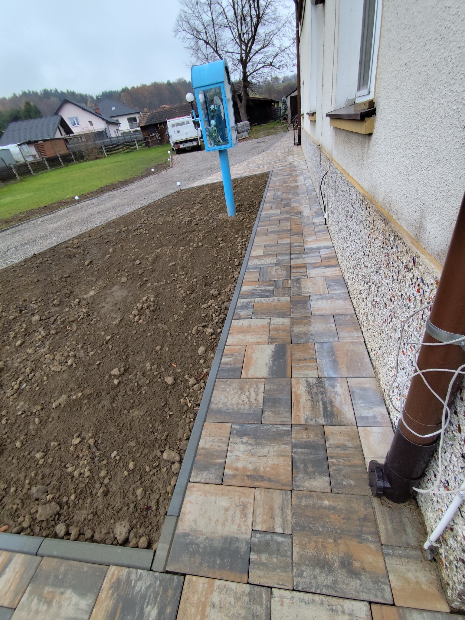 Ścieżka z kostki brukowej w Bieczu, prowadząca wzdłuż budynku. Widoczny fragment rabaty ziemnej i elementy małej architektury w tle. Kostka w odcieniach szarości i beżu.