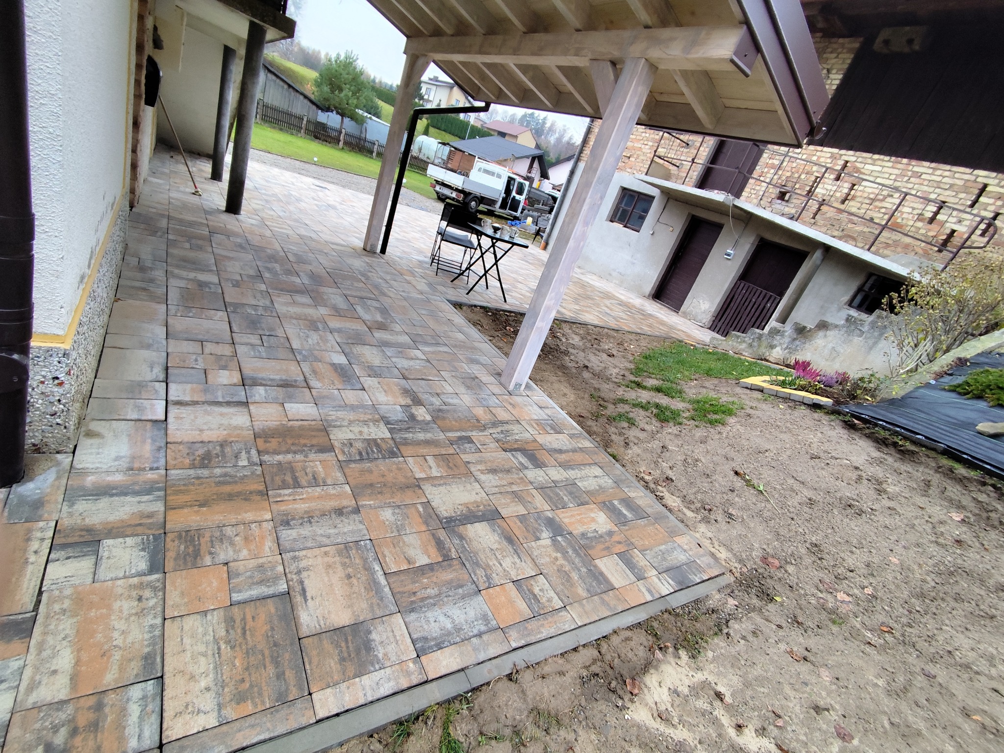 Ułożona kostka brukowa w odcieniach brązu i szarości tworzy wzór na podwórku. Widoczny fragment domu z kolumnami i stolik z krzesłami. Tło stanowi zieleń trawy i zabudowa.