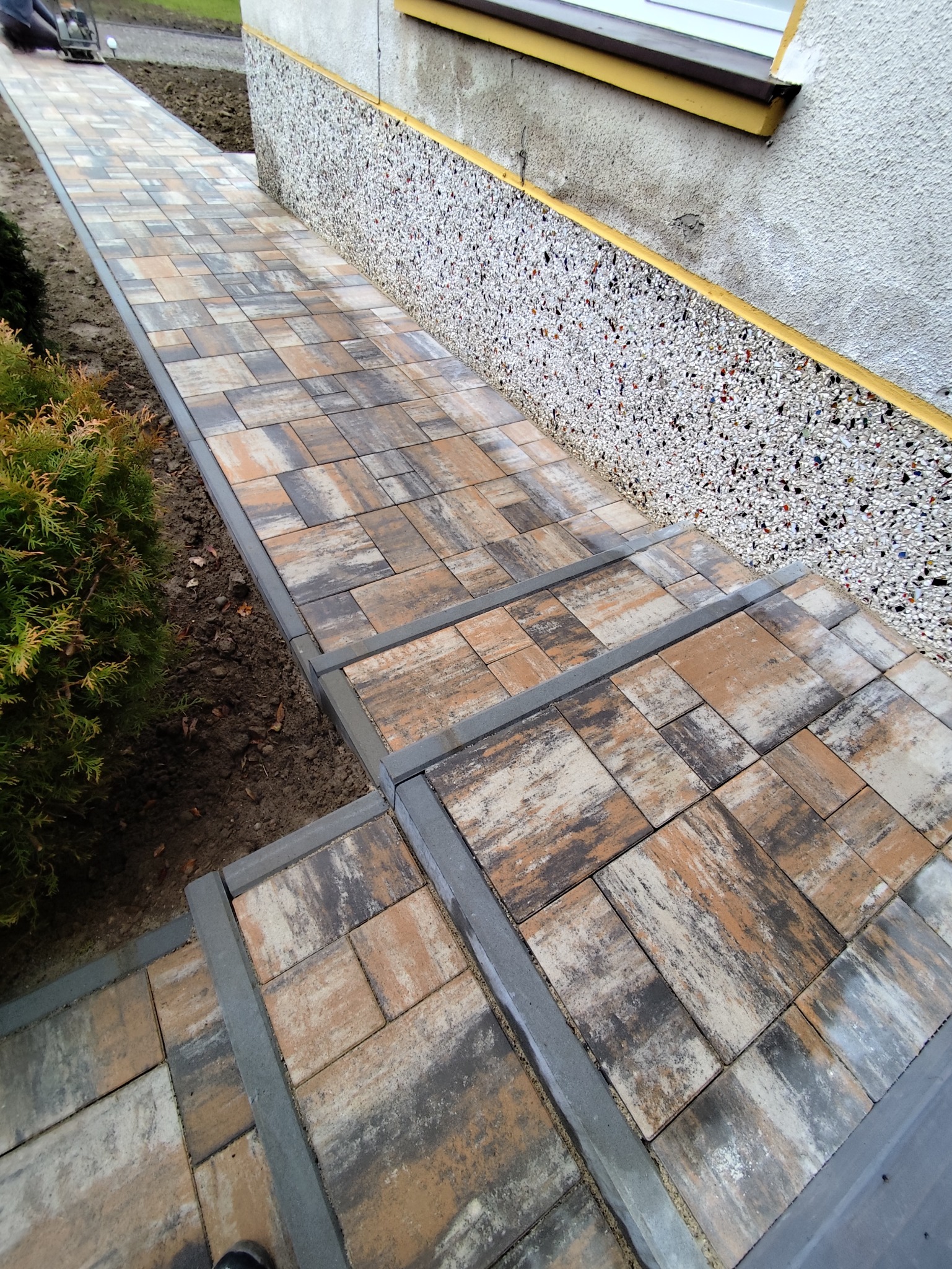 Ułożona ścieżka z kostki brukowej prowadząca do budynku z elewacją z kamyczków. Widoczne schody z kostki i trawnik z tujami. Charakterystyczny wzór kostki.