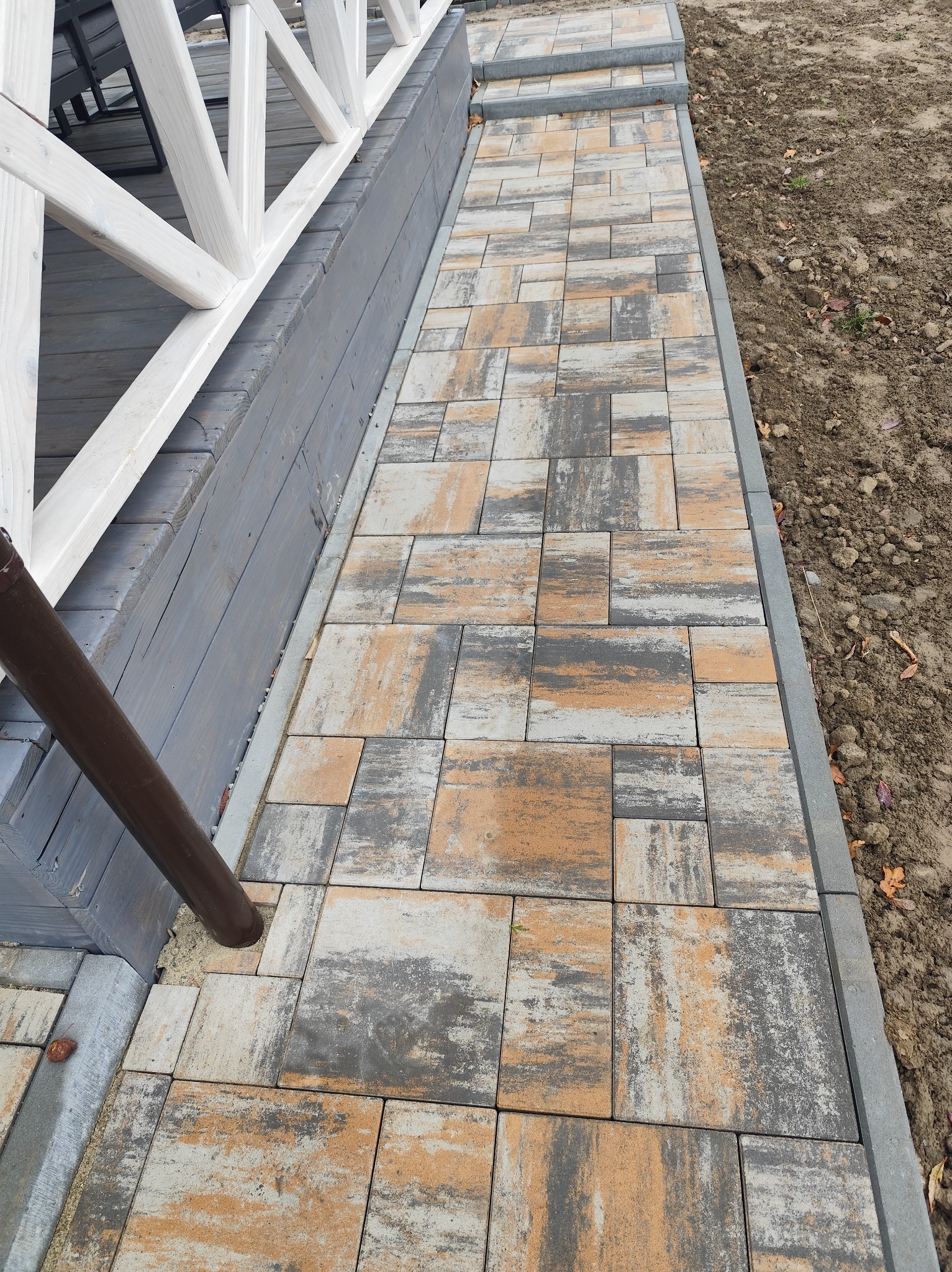 Ścieżka z kostki brukowej w odcieniach szarości i beżu, prowadząca wzdłuż drewnianej konstrukcji z białymi elementami. Widoczny brązowy spust rynnowy i fragment nieutwardzonego gruntu.