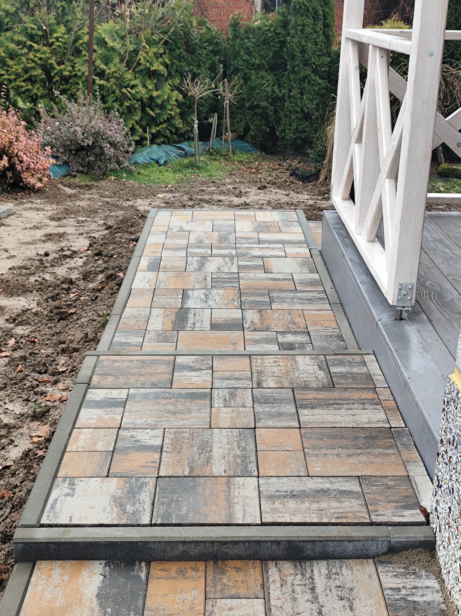 Schody z kostki brukowej o nieregularnym wzorze, prowadzące do drewnianej altany. Otoczenie w trakcie prac wykończeniowych, widoczna ziemia i roślinność.