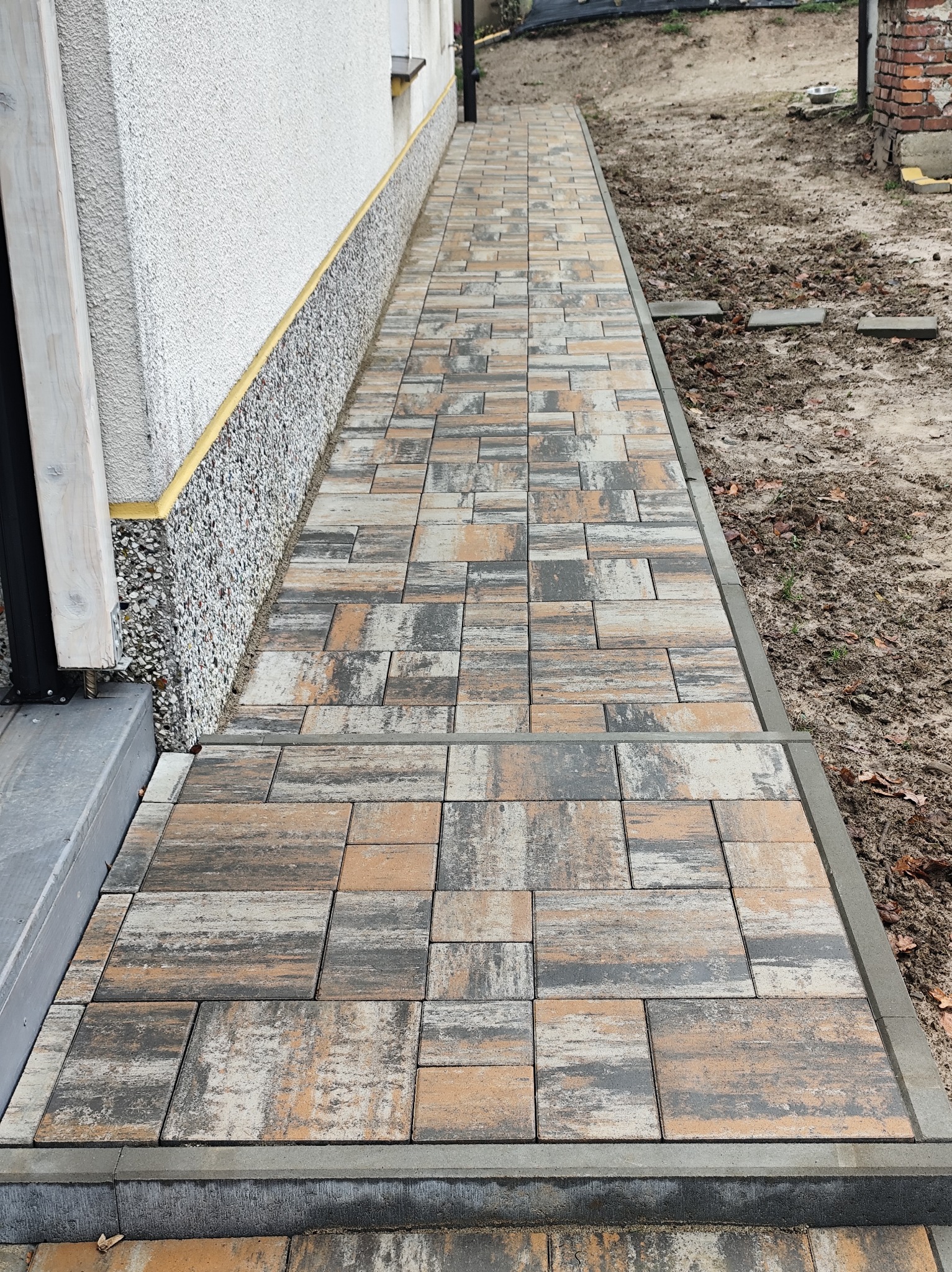 Ścieżka z kostki brukowej w odcieniach szarości i beżu, prowadząca wzdłuż ściany budynku z ozdobnym cokołem. Widoczne obrzeża i fragment terenu.