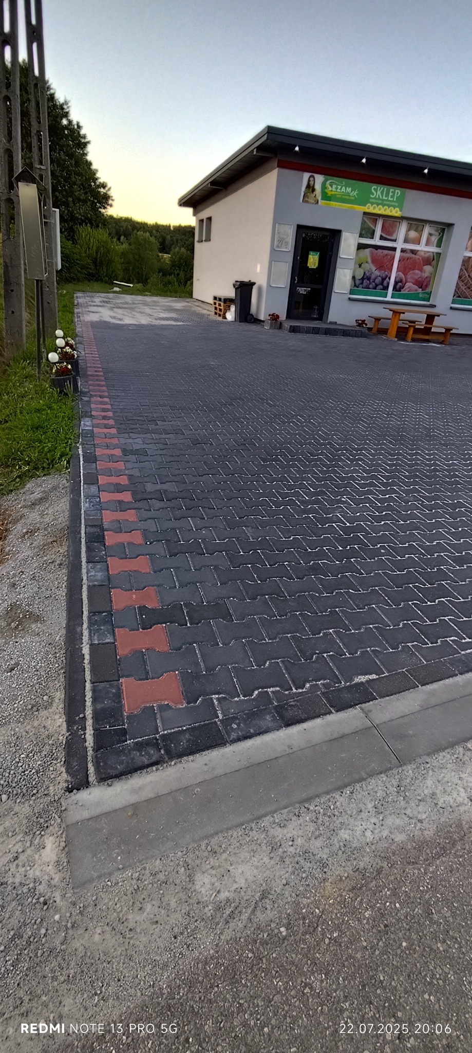 Nowa nawierzchnia z kostki brukowej w jodełkę, obrzeża z czerwonej kostki, szara kostka brzegowa. Widok na sklep z owocami i warzywami. Estetyczne wykończenie.