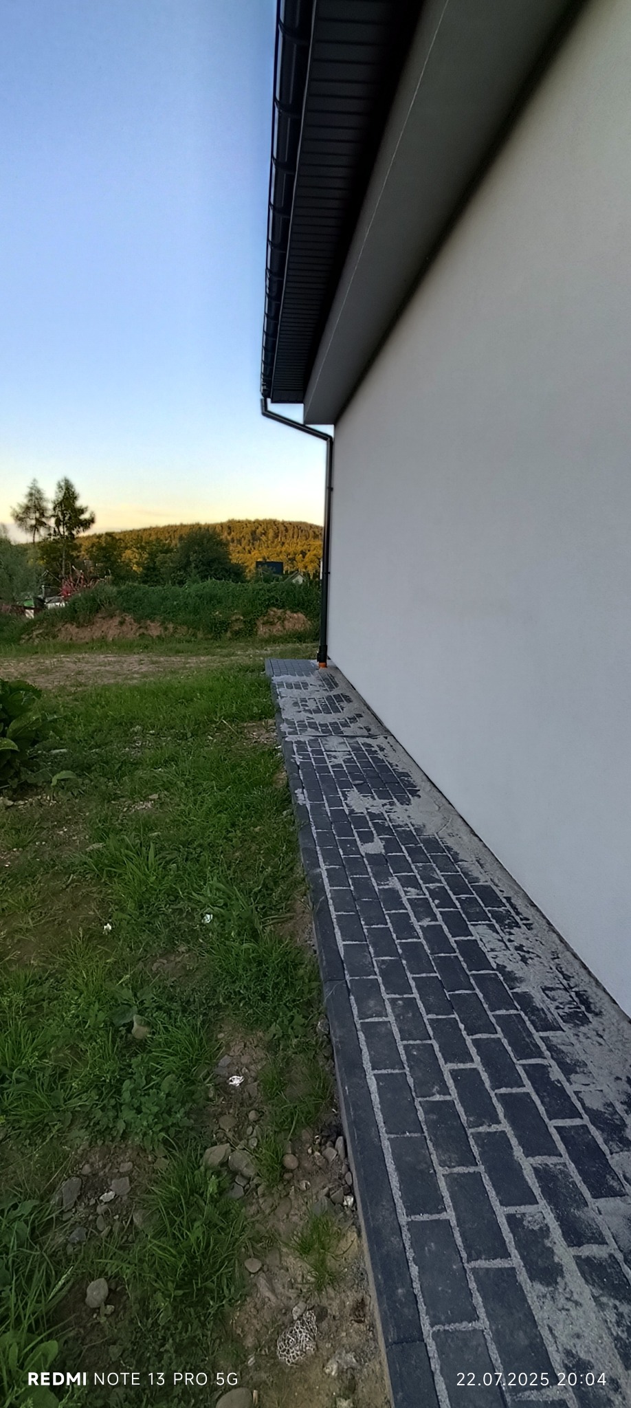 Ułożona, ciemna kostka brukowa wzdłuż białej ściany budynku, widok z boku. W tle zieleń i drzewa. Kadr z dołu, lekko zniekształcona perspektywa.