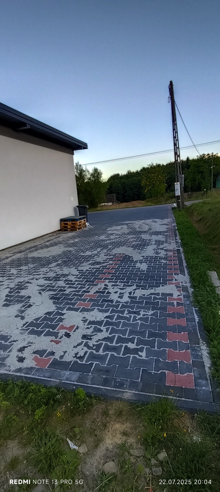 Ułożona kostka brukowa w odcieniach szarości i czerwieni, częściowo pokryta piaskiem, przylegająca do ściany budynku. Widoczne palety i słup energetyczny w tle.