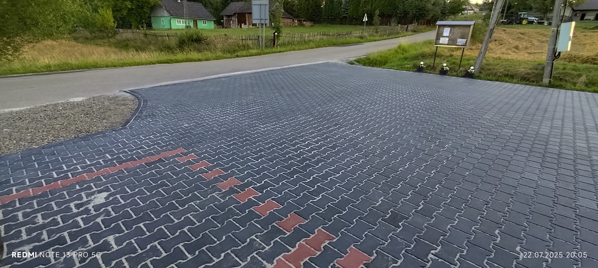 Ułożona, szara kostka brukowa z czerwonym akcentem, tworząca wzór jodełki. Widok na drogę i wiejskie zabudowania w tle. Całość w naturalnym oświetleniu.