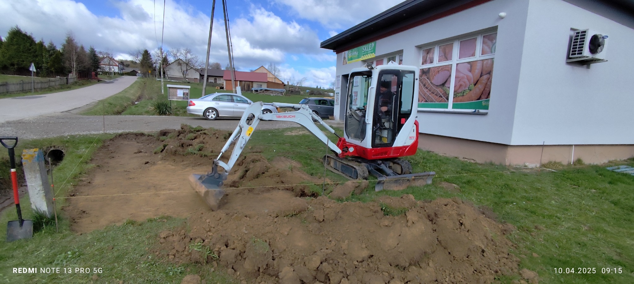 Mini koparka Takeuchi TB210 w trakcie wykopu ziemi obok budynku sklepu, widoczne narzędzia i wytyczone granice. Prace ziemne w słoneczny dzień.