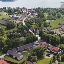 LOMA Studio - Filmy Weselne - Panoramiczny widok z lotu ptaka na wieś z czerwonymi dachami, położoną nad jeziorem, otoczoną zielonymi polami i lasami w słoneczny dzień.