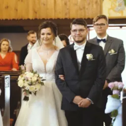 Para młoda w kościele idzie do ołtarza, w tle goście weselni, dekoracje z białych wstążek i różowych kwiatów na ławkach.