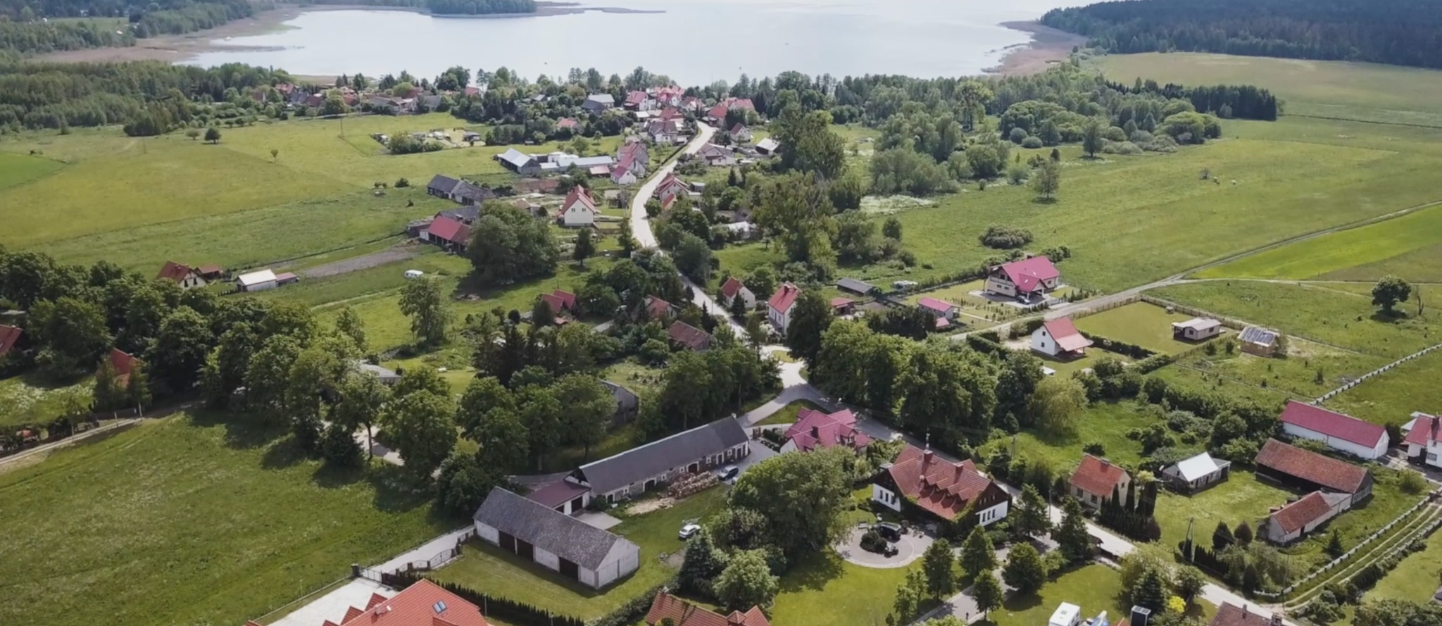 Panoramiczny widok z lotu ptaka na wieś z czerwonymi dachami, położoną nad jeziorem, otoczoną zielonymi polami i lasami w słoneczny dzień.