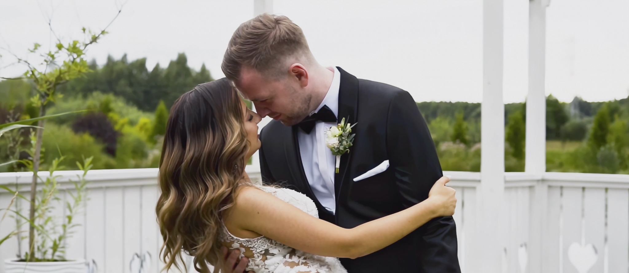 Para młoda w objęciach na tle białej altany i zieleni, panna młoda w koronkowej sukni, pan młody w czarnym smokingu z butonierką.