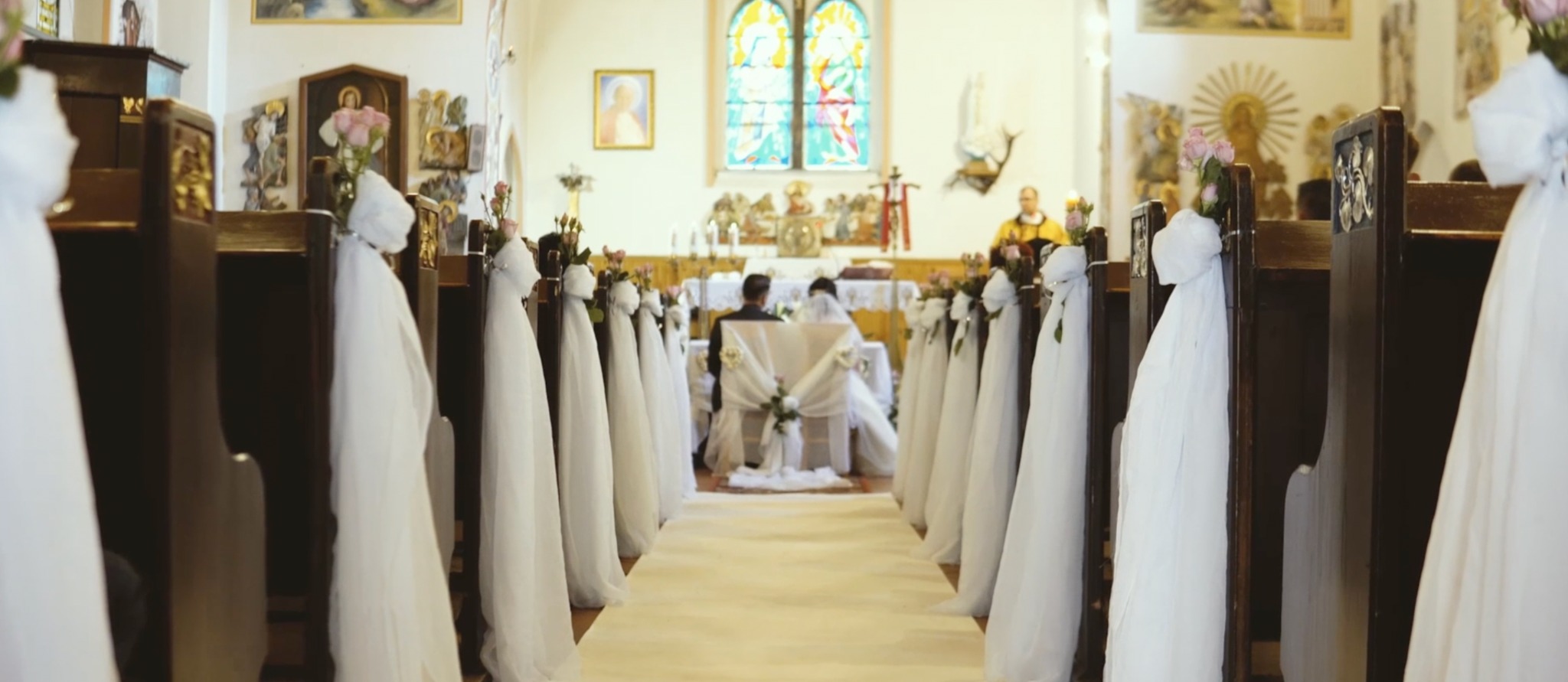 Udekorowany kościół na ślub: białe tkaniny i różowe kwiaty zdobią ławki, para młoda klęczy przed ołtarzem, ksiądz w tle.