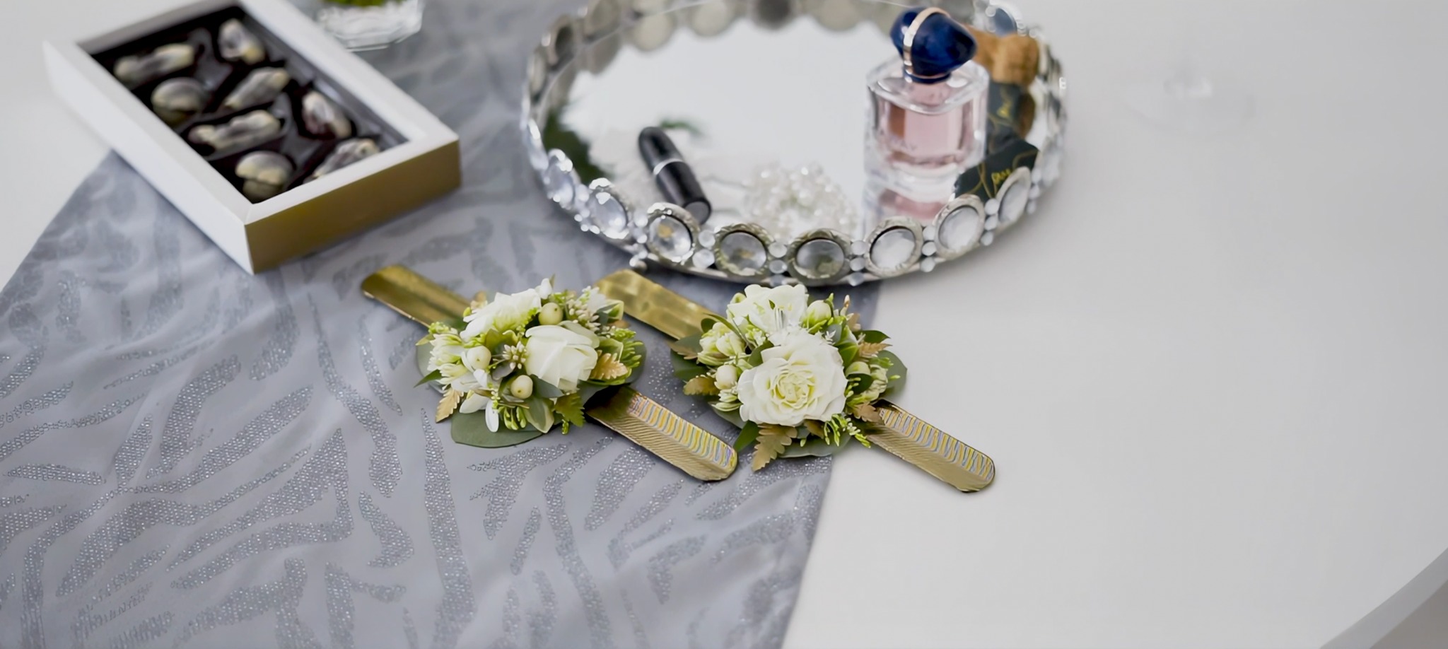 Dwa bukieciki z białych róż i zieleni na złotych podstawkach, obok pudełko czekoladek i ozdobna taca z perfumami i szminką, całość na stole z szarą i białą częścią.