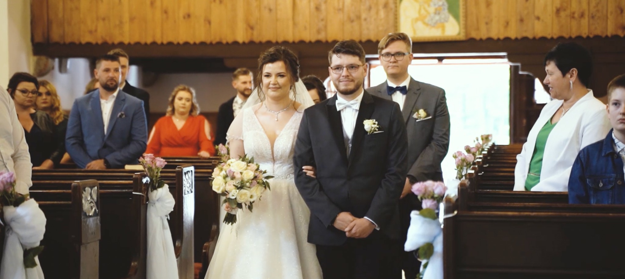 Para młoda w kościele idzie do ołtarza, w tle goście weselni, dekoracje z białych wstążek i różowych kwiatów na ławkach.
