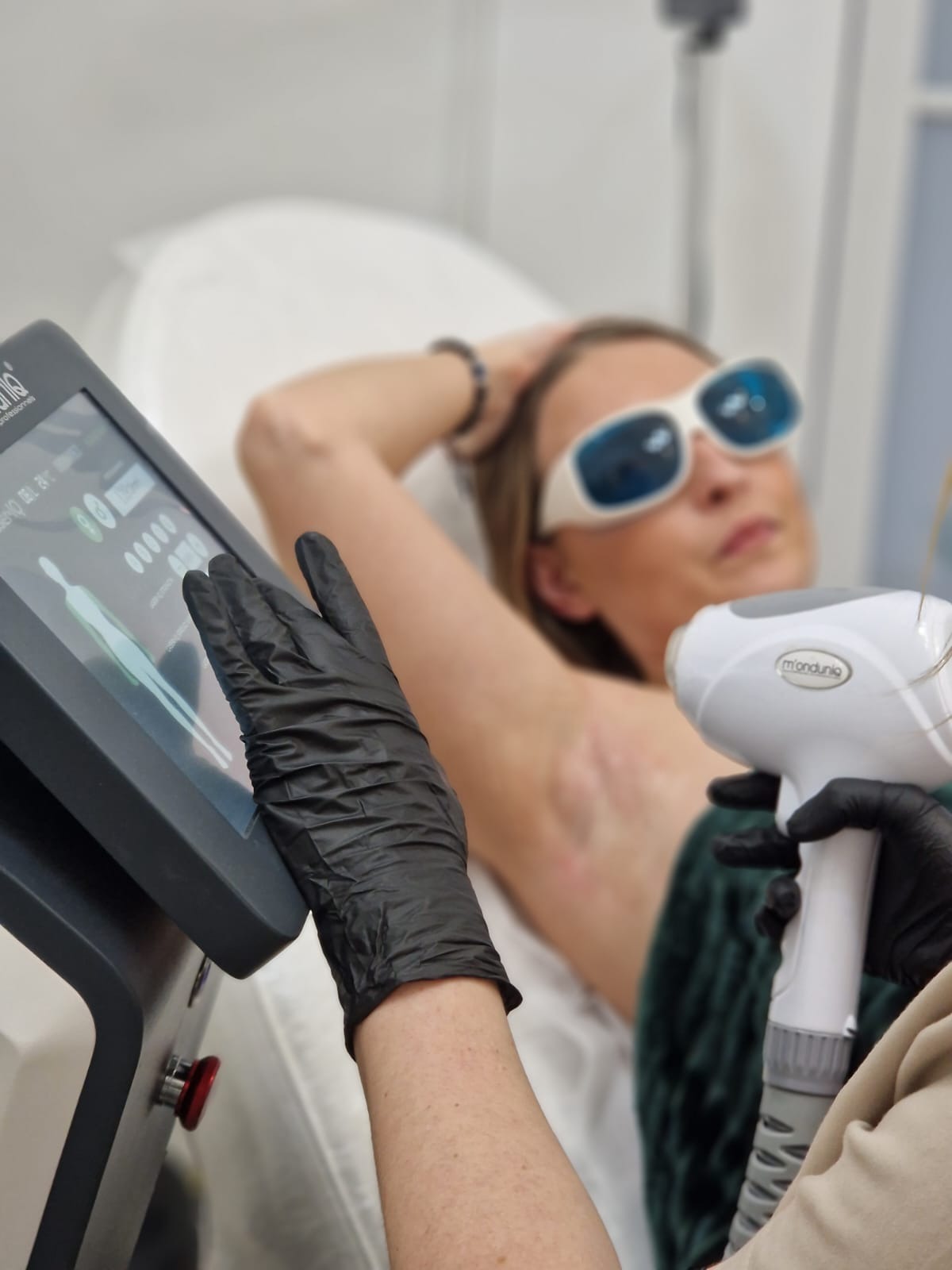 Depilacja laserowa pachy wykonywana przez osobę w czarnych rękawiczkach, z użyciem urządzenia Monduniq, pacjentka w okularach ochronnych, widoczny ekran urządzenia z ustawieniami zabiegu.