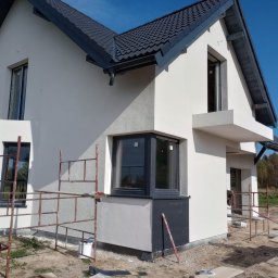 Eko - Dach - Nowoczesny dom jednorodzinny z szarym dachem i elewacją w trakcie tynkowania, widoczne rusztowania i pracownik przygotowujący płytki elewacyjne.