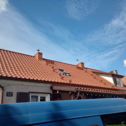 Eko - Dach - Czerwona dachówka ceramiczna na dwuspadowym dachu domu z oknem dachowym, widoczne kominy i fragment niebieskiego samochodu na pierwszym planie, błękitne niebo z chmurami w tle.