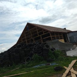 Eko - Dach - Konstrukcja dachu w trakcie prac, widoczna więźba i częściowo położone pokrycie, na pierwszym planie sterta zużytych opon, pracownik na rusztowaniu.