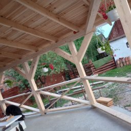 Eko - Dach - Konstrukcja drewnianej altany w trakcie budowy, widoczne elementy więźby dachowej, deski na połaci dachu, kwiaty w doniczkach wiszących pod dachem, w tle budynki gospodarcze.