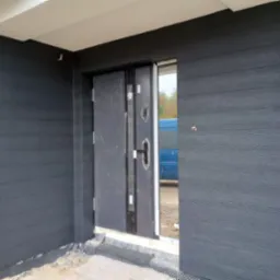 Nowoczesne wejście do domu z ciemnoszarą elewacją imitującą drewno i stalowymi drzwiami, widoczna folia fundamentowa przy podstawie elewacji.