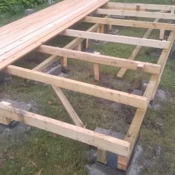 Eko - Dach - Konstrukcja drewniana pod taras, widoczne słupy na betonowych fundamentach i częściowo ułożone deski podłogowe, w tle trawnik i niebieska plandeka.