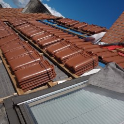 Eko - Dach - Brązowe dachówki ułożone w stosy na drewnianej konstrukcji dachu, widoczne okno dachowe i komin z cegły w tle, błękitne niebo.