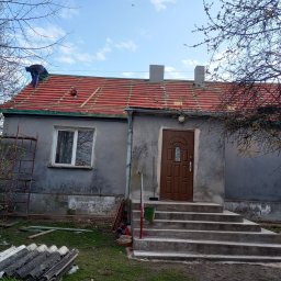 Eko - Dach - Pracownik na dachu szarego domu jednorodzinnego, montaż łat pod czerwone dachówki, widoczne rusztowanie i stos starych płyt eternitowych na trawniku.