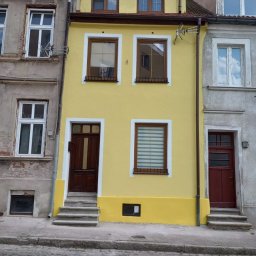 Eko - Dach - Odnowiona fasada kamienicy w kolorze żółtym, z ciemnobrązowymi drzwiami i ramami okiennymi, sąsiadująca z budynkami o elewacji szarej, widok z poziomu ulicy.