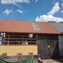Eko - Dach - Częściowo wymieniony dach z blachy trapezowej w kolorze ceglastym, z oknem dachowym na drewnianej konstrukcji budynku gospodarczego; obok fragment dachu w trakcie krycia.