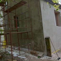 Eko - Dach - Nierównomiernie nałożony, szary tynk na ścianie budynku w trakcie prac, widoczne rusztowanie, resztki materiałów budowlanych i stara miotła oparta o betonowy słupek.