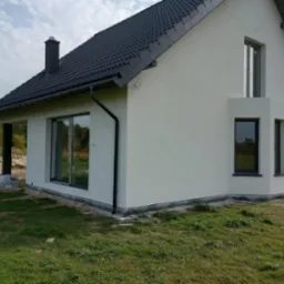 Nowoczesny dom jednorodzinny z ciemną dachówką i jasną elewacją, widoczne okna z czarnymi ramami, teren wokół domu częściowo zagospodarowany, z fragmentami trawy i ziemi.