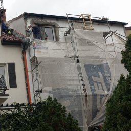 Eko - Dach - Prace elewacyjne: budynek w trakcie ocieplania, widoczne rusztowania z osłoną, pracownicy na dachu i przy oknie, cegły izolacyjne, tynk w trakcie nakładania.
