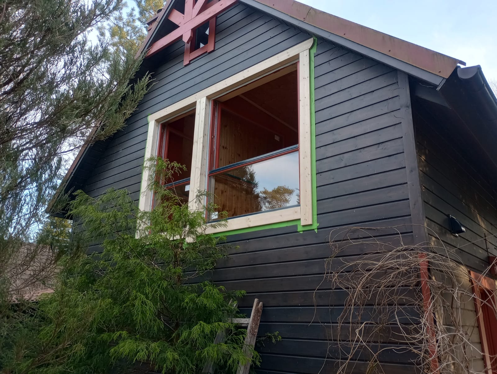 Dom z ciemną, drewnianą elewacją i nowymi oknami w trakcie montażu, zabezpieczonymi zieloną taśmą malarską. Widoczna drabina oparta o ścianę i fragment dachu z ozdobnym elementem.