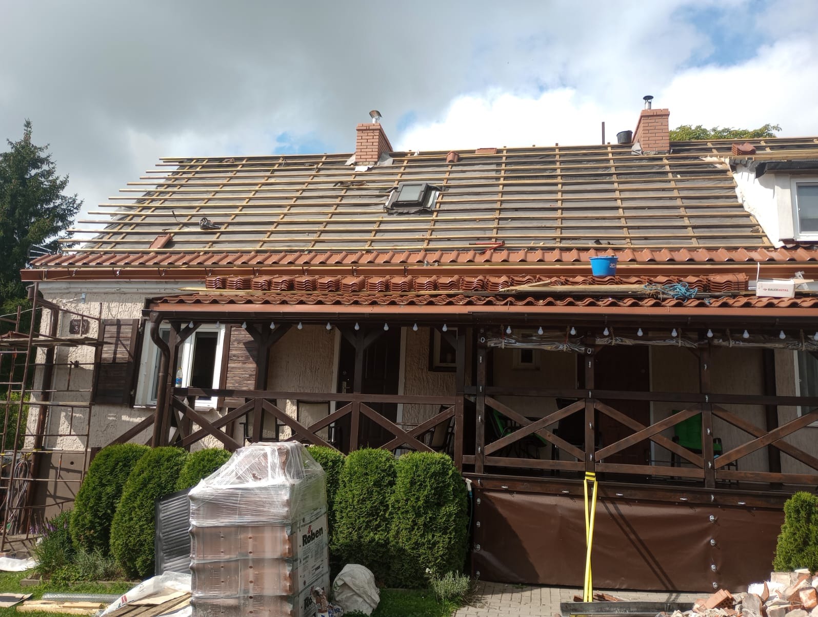 Dom w trakcie prac dekarskich, widoczne drewniane łaty na dachu, częściowo zdjęta stara dachówka i przygotowane nowe dachówki w folii na podwórku.