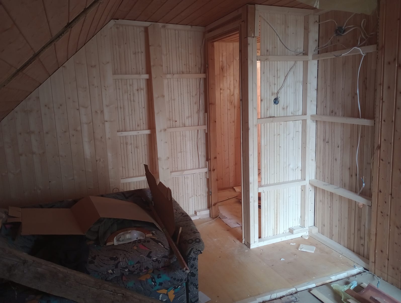 Poddasze w trakcie wykańczania boazerią, widoczne drewniane panele na ścianach, kable elektryczne, oraz fragment sofy z kartonami.