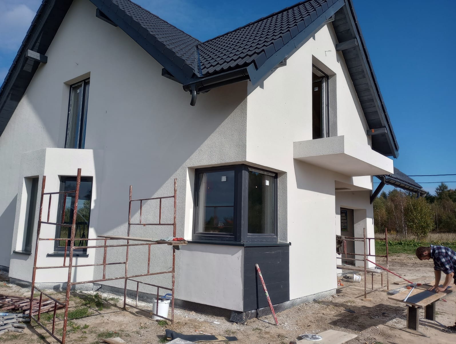 Nowoczesny dom jednorodzinny z szarym dachem i elewacją w trakcie tynkowania, widoczne rusztowania i pracownik przygotowujący płytki elewacyjne.