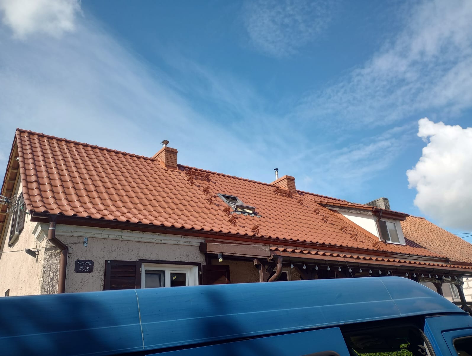 Czerwona dachówka ceramiczna na dwuspadowym dachu domu z oknem dachowym, widoczne kominy i fragment niebieskiego samochodu na pierwszym planie, błękitne niebo z chmurami w tle.