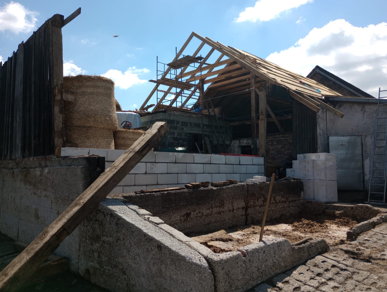 Konstrukcja drewniana dachu w trakcie budowy, widoczne rusztowania i bale siana w tle, ściany z białych bloczków.