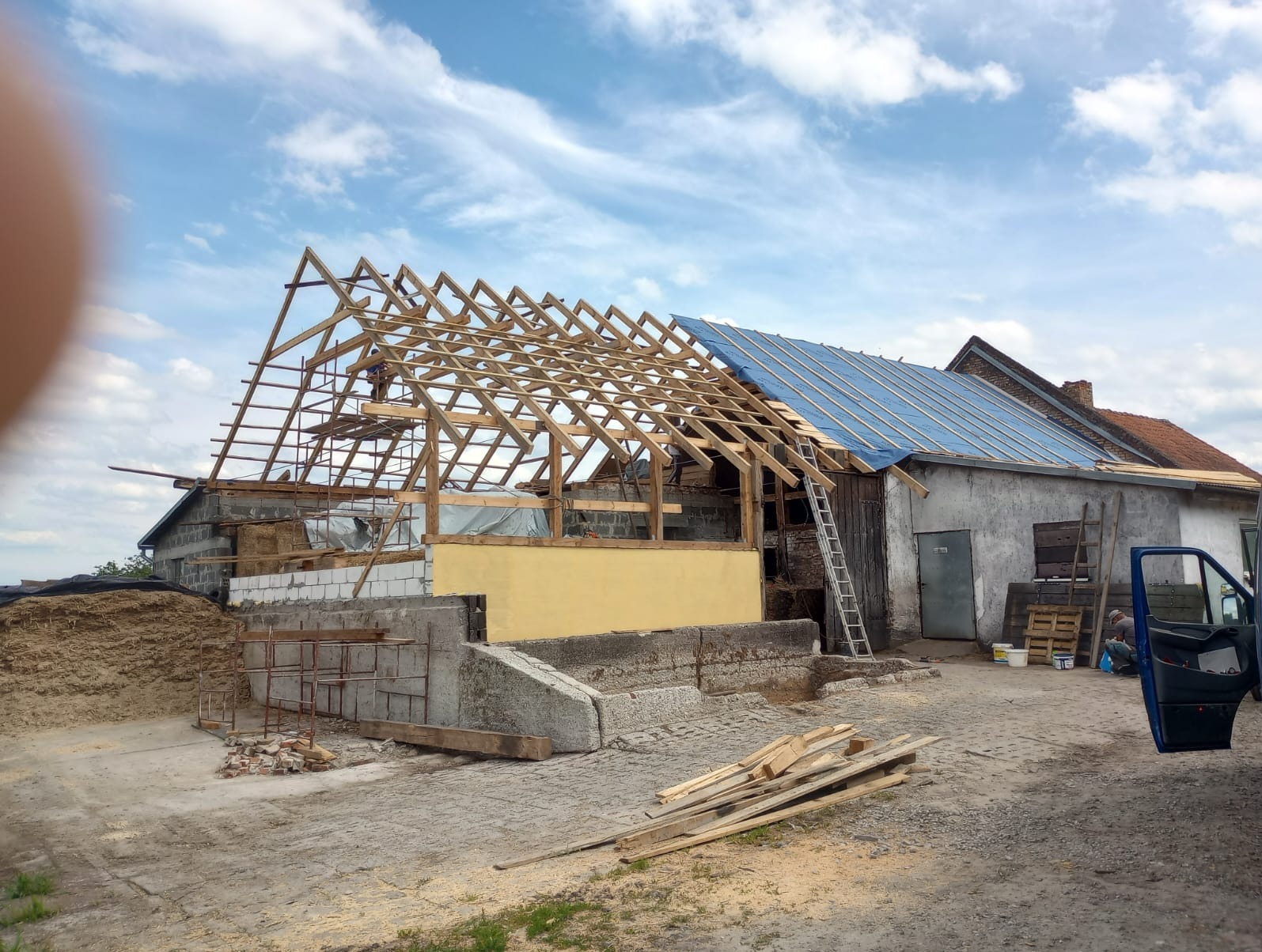Budynek gospodarczy w trakcie budowy więźby dachowej, widoczna konstrukcja drewniana oraz częściowo położona folia dachowa, w tle błękitne niebo z chmurami.