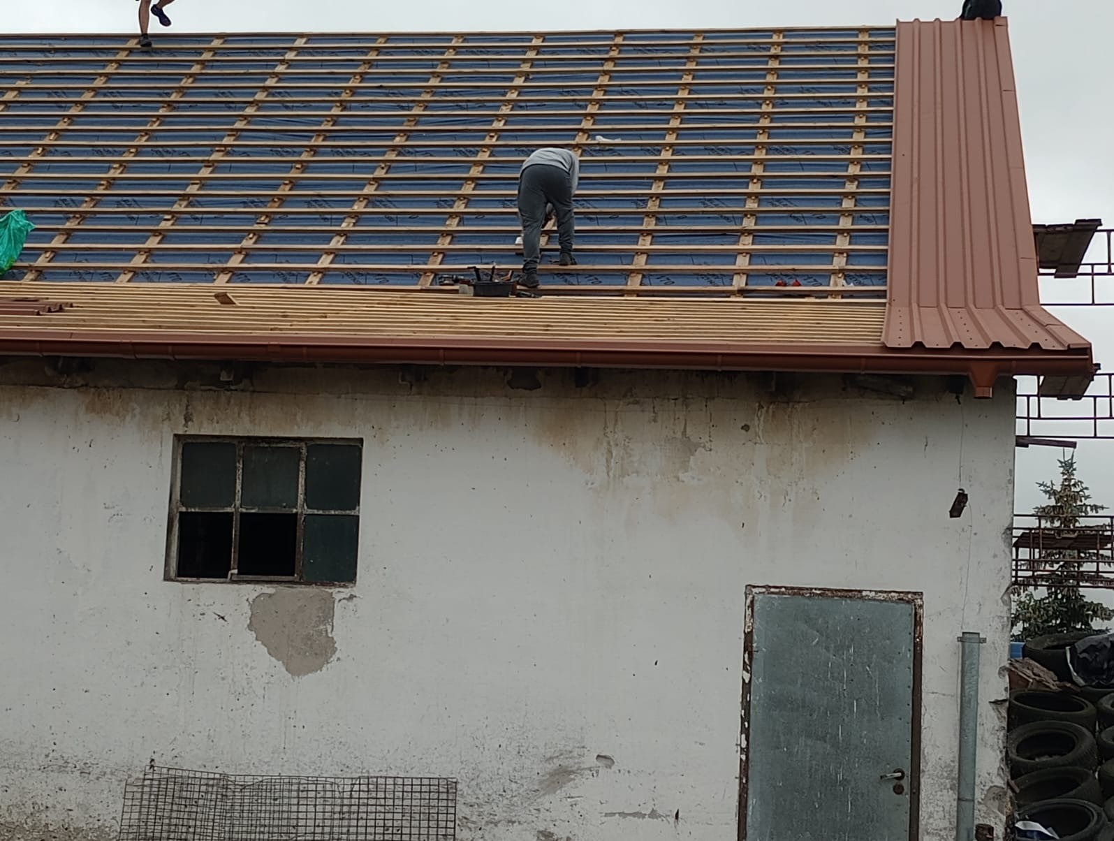 Prace na dachu budynku gospodarczego: układanie łat pod pokrycie dachowe, widoczna stara, zniszczona elewacja i okno.