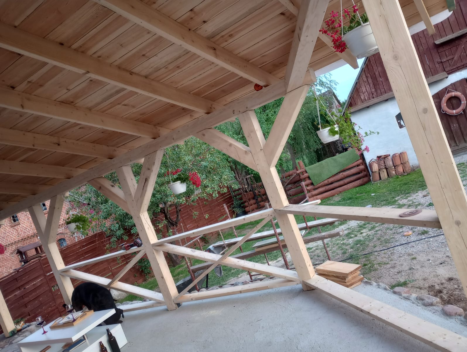 Konstrukcja drewnianej altany w trakcie budowy, widoczne elementy więźby dachowej, deski na połaci dachu, kwiaty w doniczkach wiszących pod dachem, w tle budynki gospodarcze.