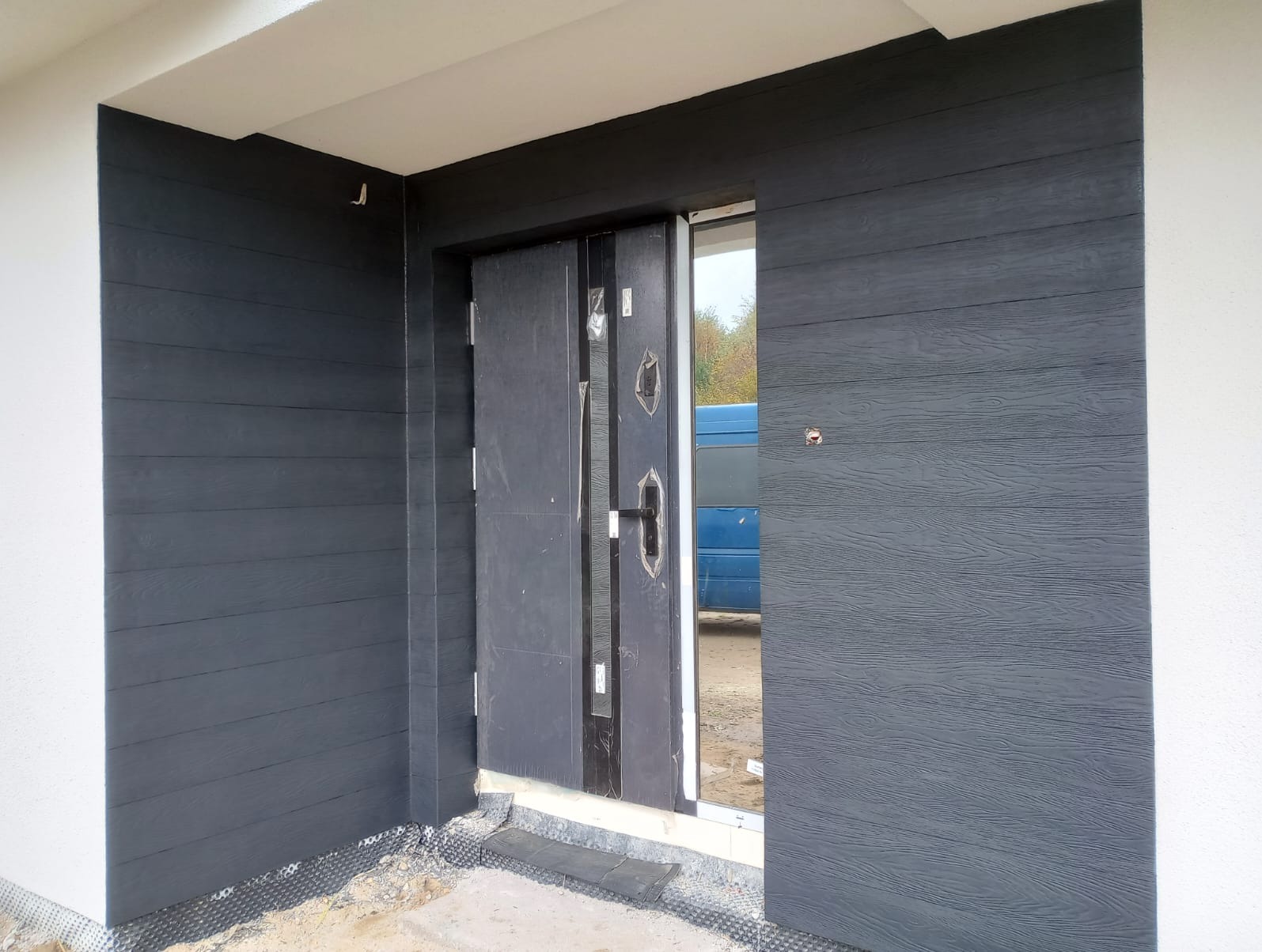 Nowoczesne wejście do domu z ciemnoszarą elewacją imitującą drewno i stalowymi drzwiami, widoczna folia fundamentowa przy podstawie elewacji.