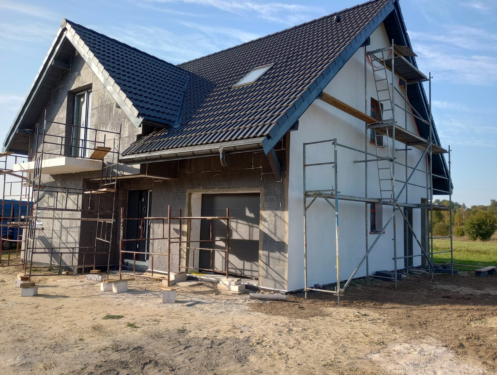 Dom w trakcie budowy: część elewacji otynkowana na biało, druga część z szarego bloczka, ciemna dachówka na dachu, rusztowania wokół budynku.