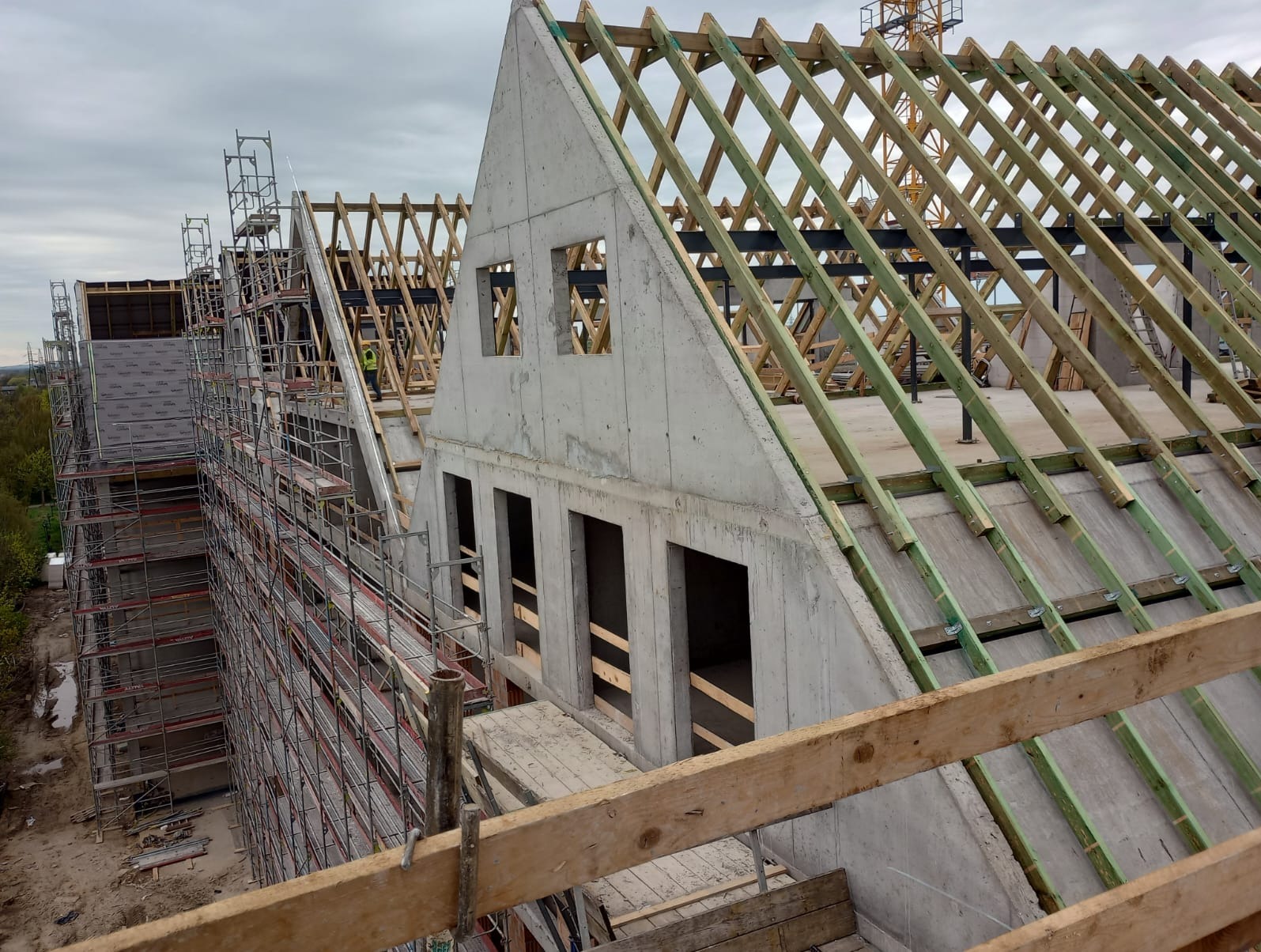 Konstrukcja dachu z betonu i drewna na budynku w trakcie budowy, widoczne rusztowania i dźwig w tle, pochmurne niebo.