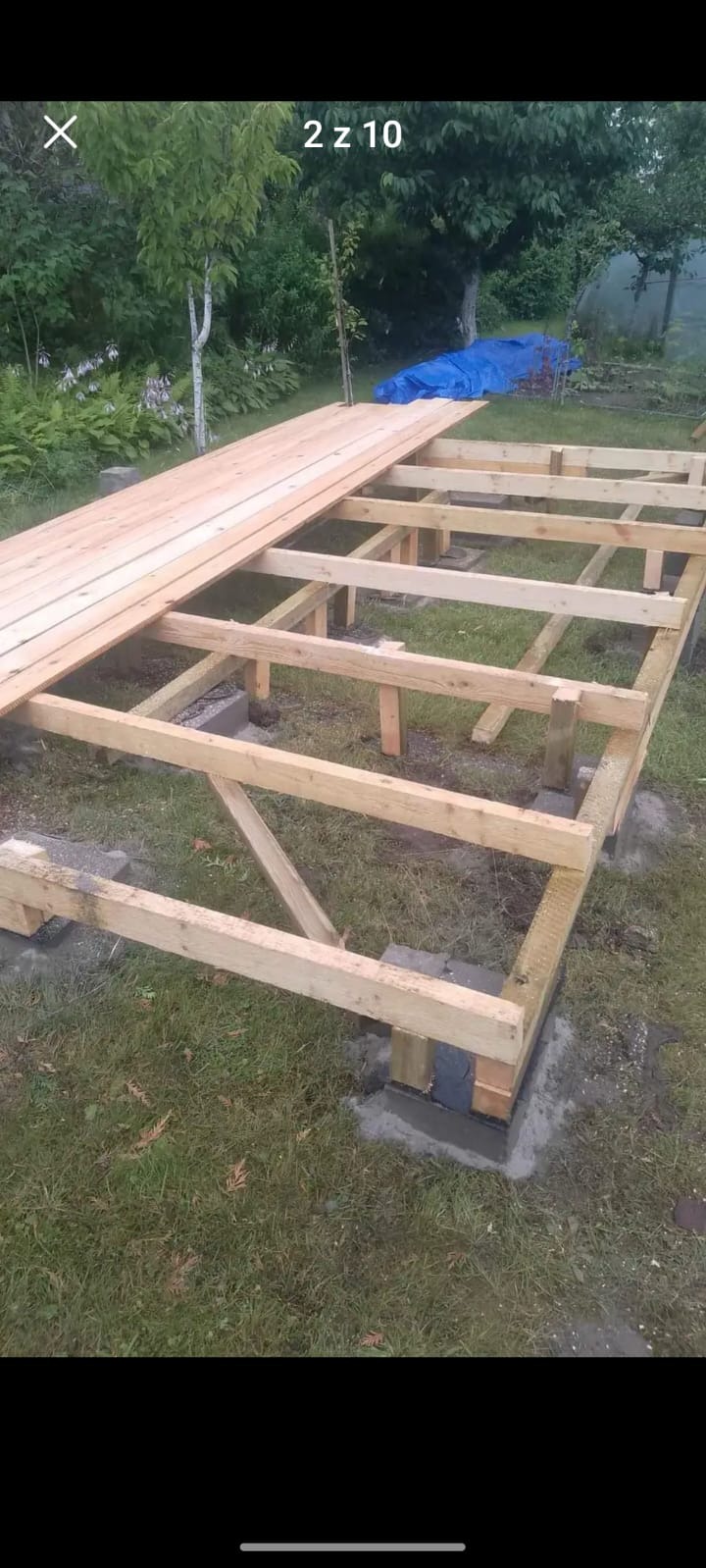 Konstrukcja drewniana pod taras, widoczne słupy na betonowych fundamentach i częściowo ułożone deski podłogowe, w tle trawnik i niebieska plandeka.