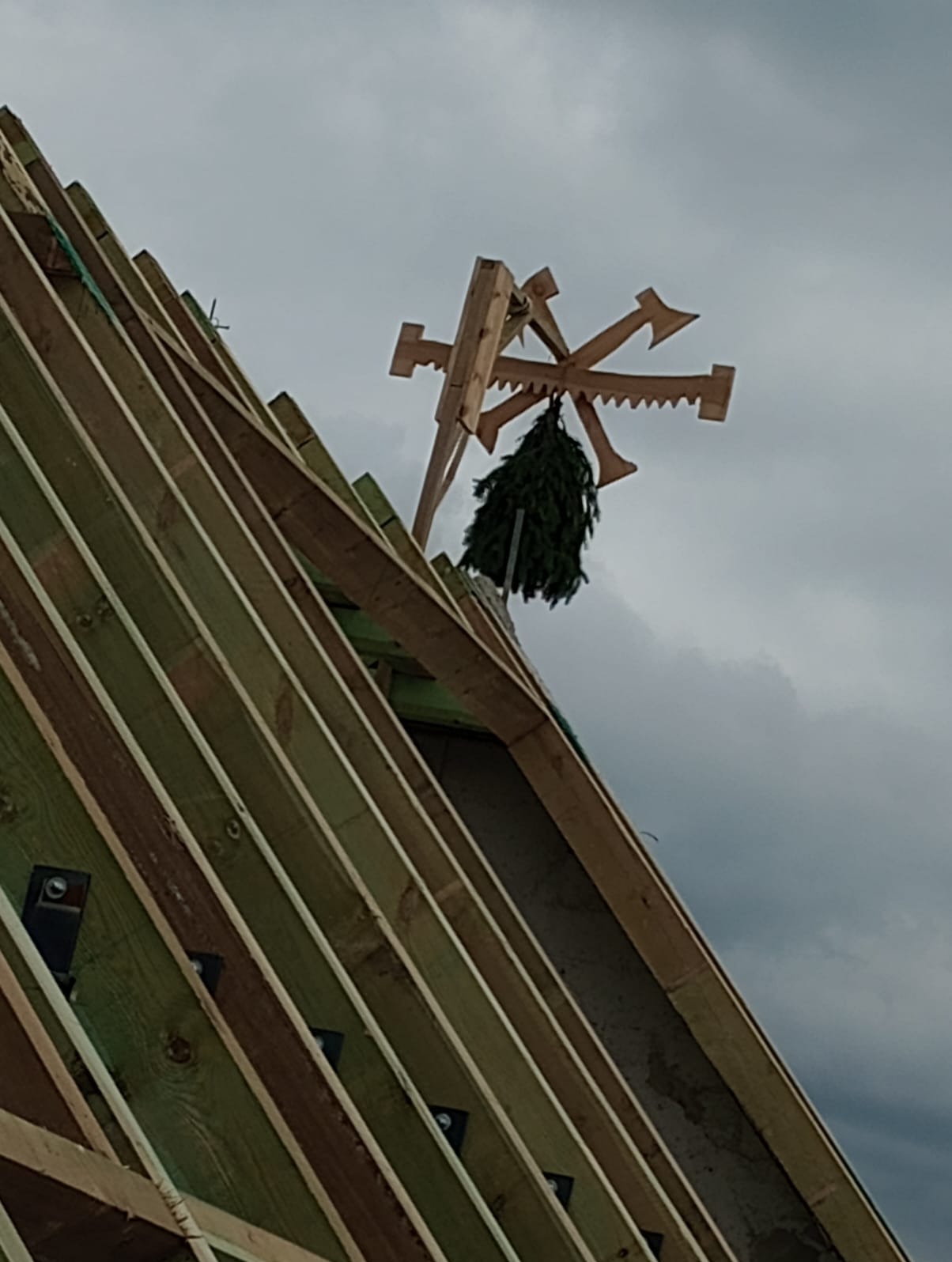 Surowa więźba dachowa z zawieszonym wieńcem na szczycie, widok z dołu na tle pochmurnego nieba.