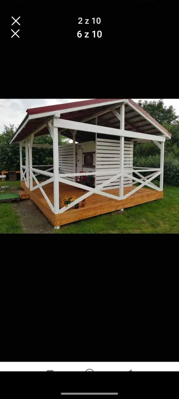 Zadaszona, drewniana altana z czerwonym dachem i białymi elementami konstrukcyjnymi, z podłogą z desek, częściowo osłaniająca przyczepę kempingową, otoczona zielenią.