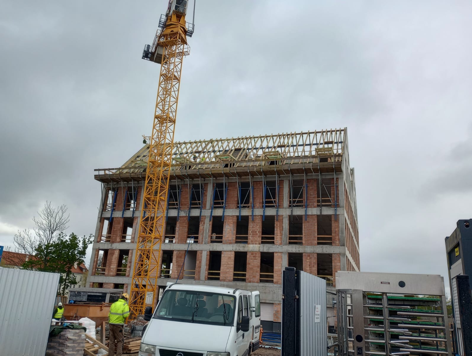 Budowa wielopiętrowego budynku z cegły, widoczna konstrukcja dachu, żółty dźwig budowlany i brygada pracowników na górze, pochmurne niebo w tle.
