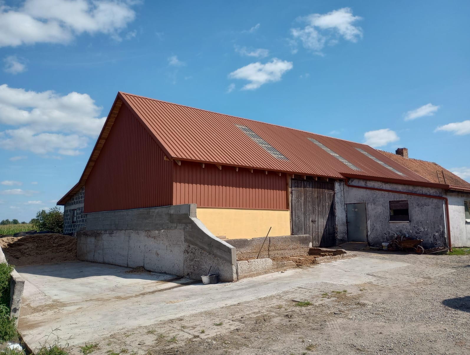 Budynek gospodarczy z nowym, brązowym dachem z blachy trapezowej i panelami ściennymi, połączony ze starszą częścią z ceglanym dachem i widocznymi śladami wilgoci na ścianach.