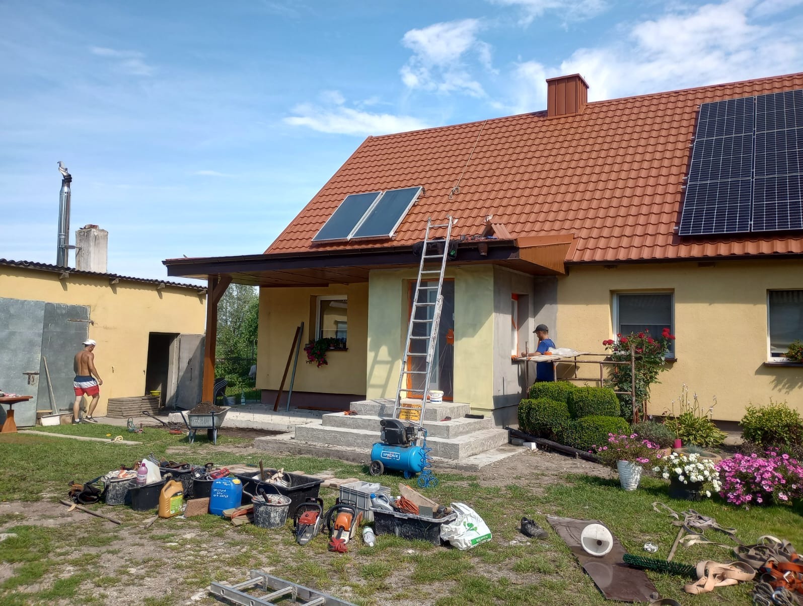 Dom w Elblągu z czerwonym dachem, na którym zamontowano panele słoneczne i kolektory; widoczna drabina oparta o okap, narzędzia i materiały budowlane na trawniku, oraz osoba pracująca przy elewacji.